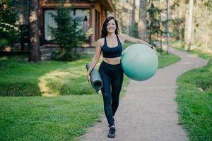 Outdoor shot of happy sporty brunette woman walks on path near forest, holds rolled up karemat and fitness ball, dressed in cropped top, leggings and sneakers, has regular trainings in open air