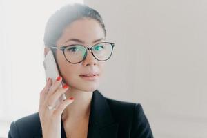 Close up shot of attractive brunette woman talks on mobile phone, looks seriously and self confident, makes call to banking service, has red manicure, wears optical glasses, formal black suit photo