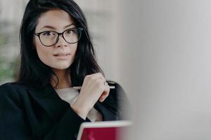 Self confident serious adult female employee looks though transparent glasses, writes information while working on project, makes plans, dressed in formal wear, notes some records in notepad photo