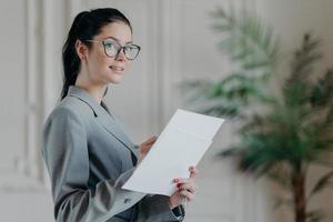 Successful brunette businesswoman stands thoughtful with paper documents, busy with making startup, analyzes business information, writes down notes, looks somewhere into distance, dressed formally photo