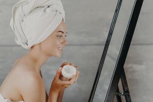 una foto lateral de una mujer feliz renovada posa con los hombros desnudos, usa una toalla de baño en la cabeza, se mira en el espejo, se aplica crema facial, tiene una piel sana, usa cosméticos naturales. concepto de autocuidado