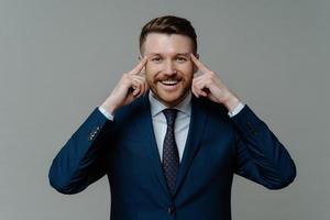 Cheerful businessman in suit holding fingers on temples and smiling at camera photo