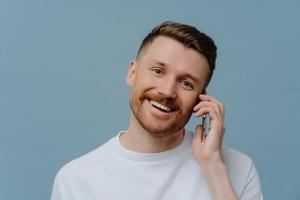 Indoor shot of handsome bearded man enjoys telephone talk smiles toothily dressed in casual white t shirt isolated over blue background. Unshaven happy guy discusses news with friend. Communication photo