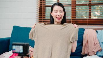 Asian stylist fashion influencer designer women using camera streaming and live to sell clothes, product, and accessories business, e-commerce broadcast online on sofa in living room at home. photo