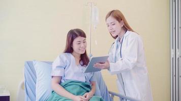 Beautiful smart Asian doctor and patient discussing and explaining something with tablet in doctor hands while staying on Patient's bed at hospital. Medicine and health care concept. photo