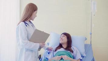 Beautiful smart Asian doctor and patient discussing and explaining something with clipboard in doctor hands while staying on Patient's bed at hospital. Medicine and health care concept. photo