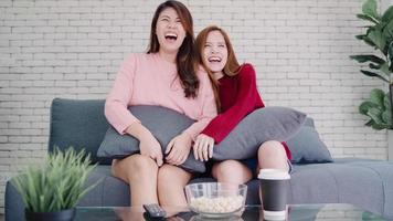 Lesbian Asian couple watching TV laugh and eating popcorn in living room at home, sweet couple enjoy funny moment while lying on the sofa when relaxed at home. Lifestyle couple relax at home concept. photo