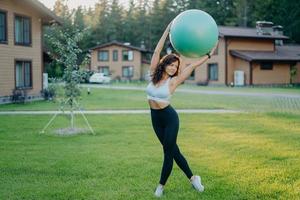 Full length shot of serious slim brunette woman has perfect body shape holds fitness ball over head, does fitness exercises outdoor near house, dressed in cropped top, leggings and sneakers. photo