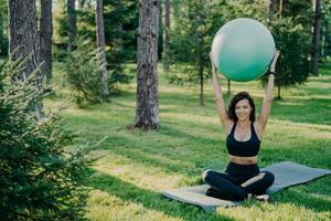 Brunette slim woman lifts fitness ball over head, sits in lotus pose on karemat, dressed in active wear, practices yoga outside, breathes fresh air in forest. Fit lady exercises with gymnastic ball photo