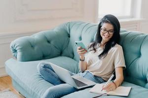 Indoor shot of happy female freelancer holds mobile phone, makes necessary notes in personal diary, uses modern technologies for sharing media, sits on comfortable couch in spacious living room