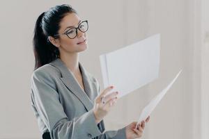 Photo of female financial analyst makes report with recommendations for investments, reads strategy information, poses with paper documents, dressed in formal clothes, wears eyeglasses, stands indoor