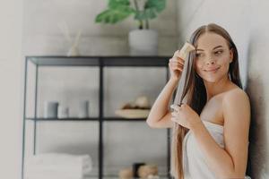Attractive young woman smiles and enjoys her beauty, combes long straight hair after applying mask, stands wrapped in bath towel in cozy bathroom, has smooth skin. Hair care, spa and beauty concept photo