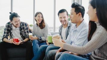 diversidad del equipo del grupo de jóvenes sosteniendo tazas de café y discutiendo algo con una sonrisa mientras está sentado en el sofá de la oficina. tiempo de descanso para tomar café en la oficina creativa. foto