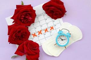 Menstrual pads on menstruation period calendar with blue alarm clock and red rose flowers photo