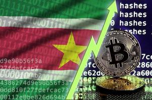 bandera de surinam y flecha verde ascendente en la pantalla de minería bitcoin y dos bitcoins dorados físicos foto