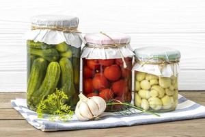 Homemade pickled cherry tomatoes, cucumbers, champignons, garlic, eggplant, red peppers in jars on wooden shelf Homemade canned and fermented foods concept Seasonal product photo