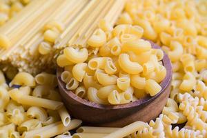 macarrones crudos en tazón de madera, pasta varios tipos de espaguetis de pasta sin cocer y fideos en el fondo, concepto culinario de comida italiana, colección de diferentes pastas crudas para cocinar alimentos foto