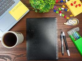 endecha plana, maqueta de libro negro. espacio de trabajo en el fondo del escritorio de la oficina desde la vista superior. con libros negros, computadoras portátiles, material de oficina, lápices, hojas verdes y tazas de café en la mesa de madera. foto