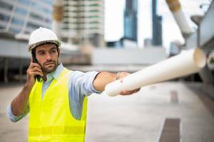 el ingeniero caucásico usa un teléfono inteligente para hablar, usa chaleco amarillo y un gran sombrero duro, y la otra mano sostiene el plano de piso blanco en el sitio de trabajo del centro de la ciudad. foto