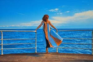 Pretty girl in flowing long dress stands on edge of pier. photo