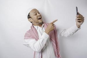 A portrait of a happy Asian Muslim man smiling while holding his phone, isolated by white background photo