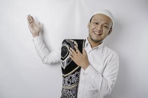 Smiling Asian Muslim man with prayer rug on his shoulder is pointing at the copy space on top of him, isolated by white background photo