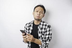 A dissatisfied young Asian man looks disgruntled wearing tartan shirt irritated face expressions holding his phone photo