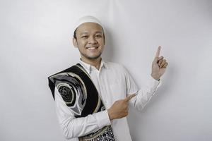 Smiling Asian Muslim man with prayer rug on his shoulder is pointing at the copy space on top of him, isolated by white background photo