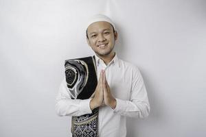 Smiling young Asian Muslim man with prayer rug on his shoulder, gesturing traditional greeting isolated over white background photo