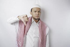 Disappointed Asian Muslim man gives thumbs down hand gesture of approval, isolated by white background photo