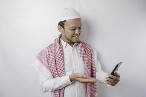 A portrait of a happy Asian Muslim man smiling while holding his phone, isolated by white background photo