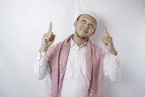 Smiling Asian Muslim man pointing at the copy space on top of him, isolated by white background photo