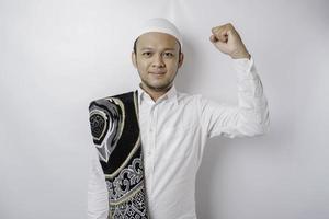 A happy young Asian Muslim man with a prayer rug on his shoulder showing a successful expression isolated by white background photo