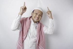 Smiling Asian Muslim man pointing at the copy space on top of him, isolated by white background photo
