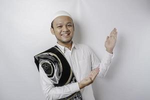 Smiling Asian Muslim man with prayer rug on his shoulder is pointing at the copy space on top of him, isolated by white background photo