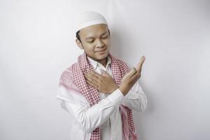 Smiling Asian Muslim man pointing at the copy space on top of him, isolated by white background photo