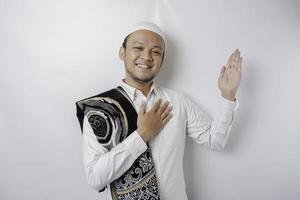 Smiling Asian Muslim man with prayer rug on his shoulder is pointing at the copy space on top of him, isolated by white background photo