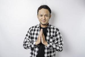 Smiling young Asian man wearing tartan shirt, gesturing traditional greeting isolated over white background photo