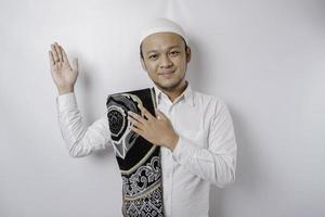Smiling Asian Muslim man with prayer rug on his shoulder is pointing at the copy space on top of him, isolated by white background photo