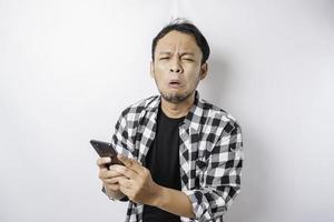 A dissatisfied young Asian man looks disgruntled wearing tartan shirt irritated face expressions holding his phone photo