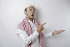 Shocked Asian Muslim man pointing at the copy space beside him, isolated by white background photo