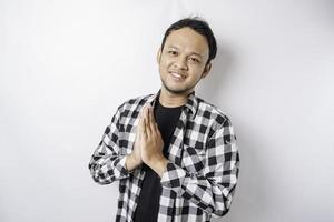 Smiling young Asian man wearing tartan shirt, gesturing traditional greeting isolated over white background photo