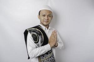 Smiling young Asian Muslim man with prayer rug on his shoulder, gesturing traditional greeting isolated over white background photo