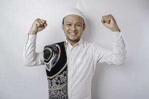 A happy young Asian Muslim man with a prayer rug on his shoulder showing a successful expression isolated by white background photo