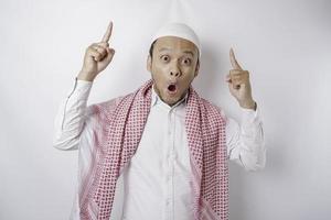 Shocked Asian Muslim man pointing at the copy space on top of him, isolated by a white background photo