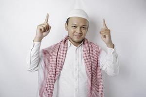 Smiling Asian Muslim man pointing at the copy space on top of him, isolated by white background photo