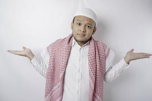 A thoughtful young Muslim man shrugging his shoulders, gesturing confusion isolated by white background photo