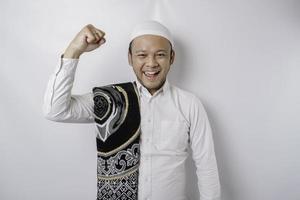 A happy young Asian Muslim man with a prayer rug on his shoulder showing a successful expression isolated by white background photo