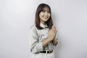 una joven asiática sonriente con pantalones verde salvia, haciendo un gesto de saludo tradicional aislada sobre fondo blanco foto