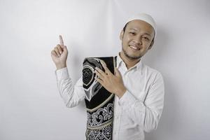 Smiling Asian Muslim man with prayer rug on his shoulder is pointing at the copy space on top of him, isolated by white background photo